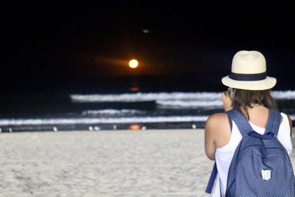 chica morena de espaldas con un sombrero y fotografiando la noche en una playa